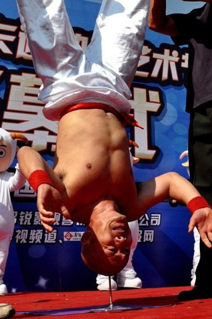 Китаец Ли Синь выполняет немыслимый трюк — он стоит вверх ногами, опираясь головой на острие гвоздя, причём без помощи рук. 